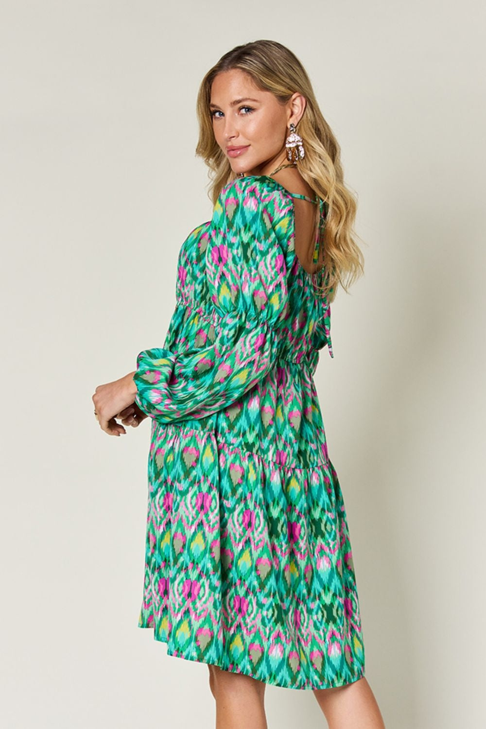 A model looks over her shoulder in a vibrant green and pink printed long sleeve dress with ruched waist detailing and a flowing A-line skirt.