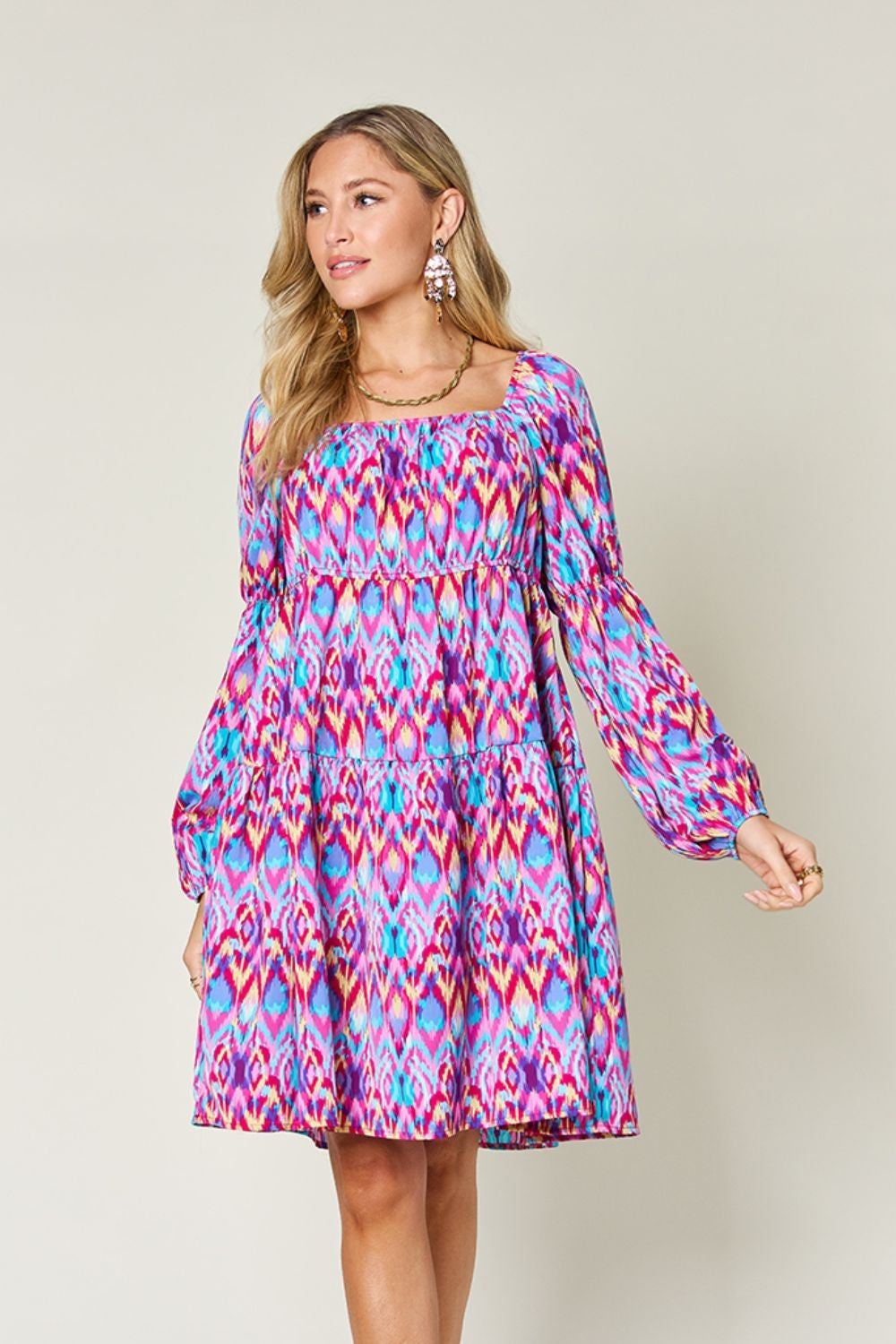 Model poses in a vibrant pink, blue, and purple printed long sleeve dress with a square neckline, ruched waist, tied detail, tiered construction, and easy A-line silhouette.