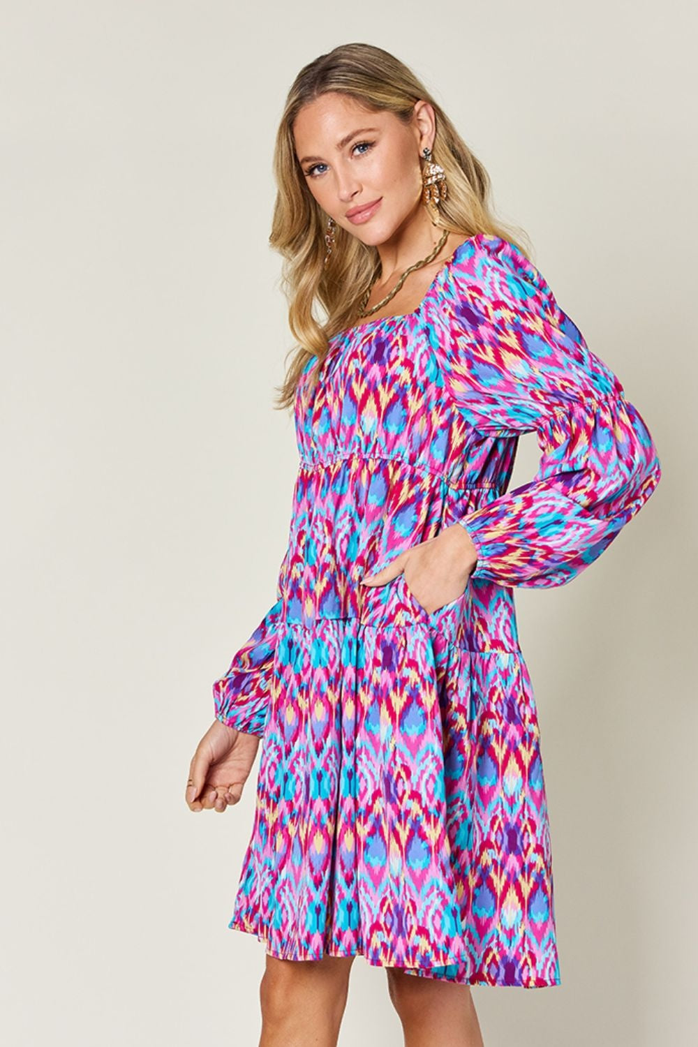 Model poses with one hand on her hip in a vibrant pink, turquoise, and blue printed long sleeve dress with a ruched waist and flowing A-line skirt.
