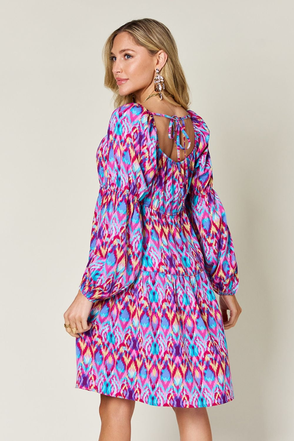 Model turns to show a vibrant printed long sleeve dress with ruched waist, tiered A-line silhouette, voluminous sleeves, and tied open back.