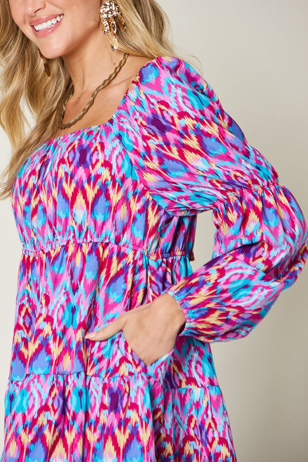 A model poses with one hand on her hip in a vibrant printed long sleeve dress featuring a rounded neckline, ruched waist, tied detail, and flowing A-line silhouette.