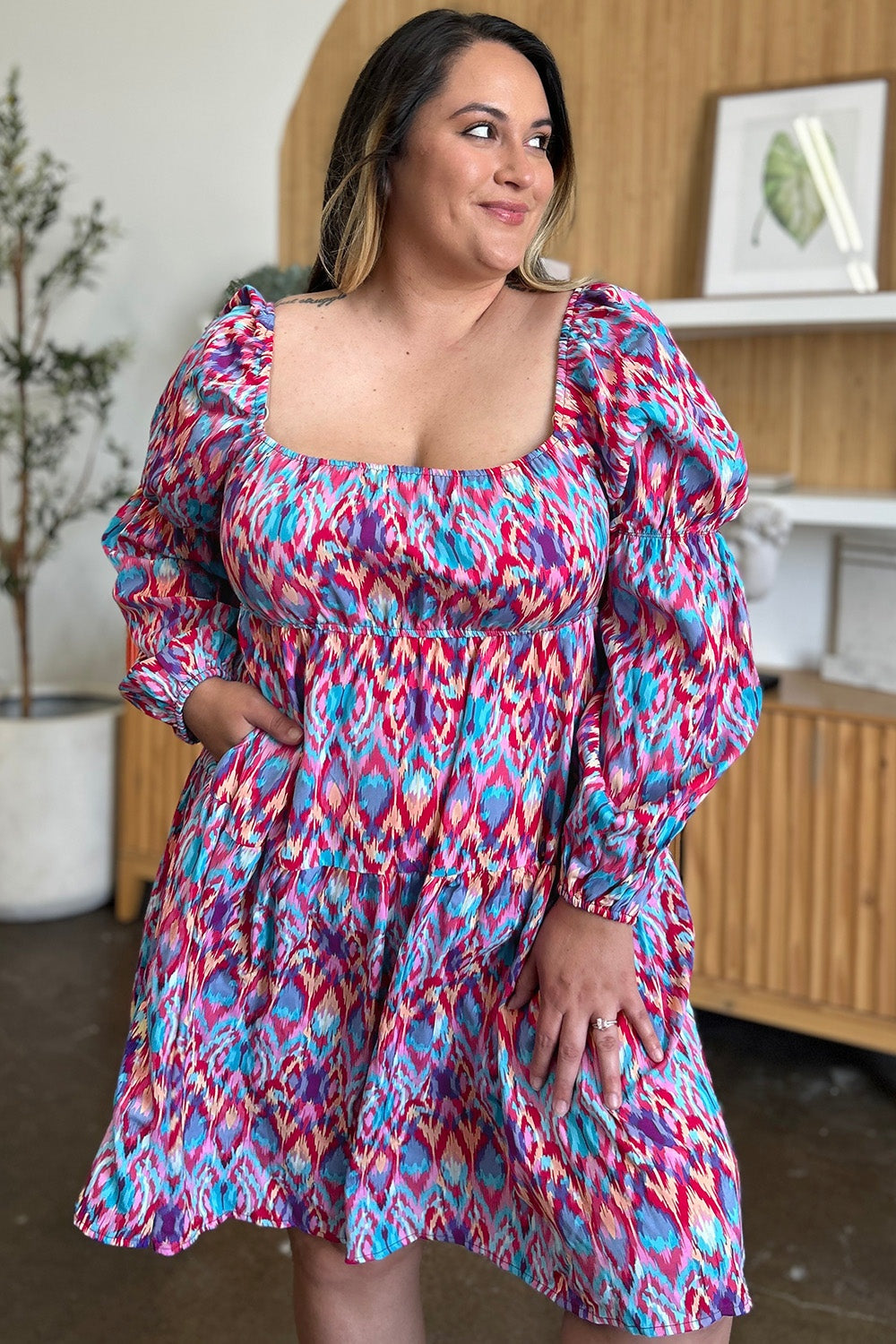 A model wears a vibrant blue, pink, and red printed long sleeve dress with a square neckline, gathered waist, puff sleeves, elastic cuffs, and a flowing A-line skirt.