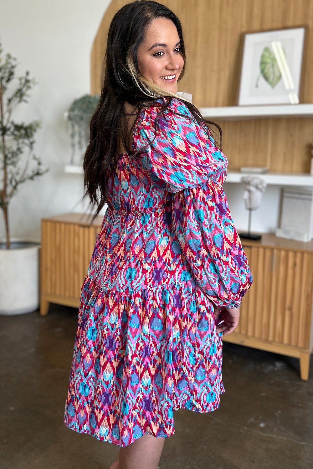 Model turns to show a vibrant printed long sleeve dress with ruched waist, gathered sleeves, tied neckline, and flowing A-line skirt.