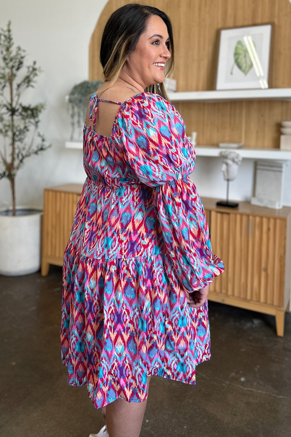 A model turns to display a vibrant red, blue, and pink printed long sleeve dress with a tied back neckline, ruched waist, and gathered A-line skirt.