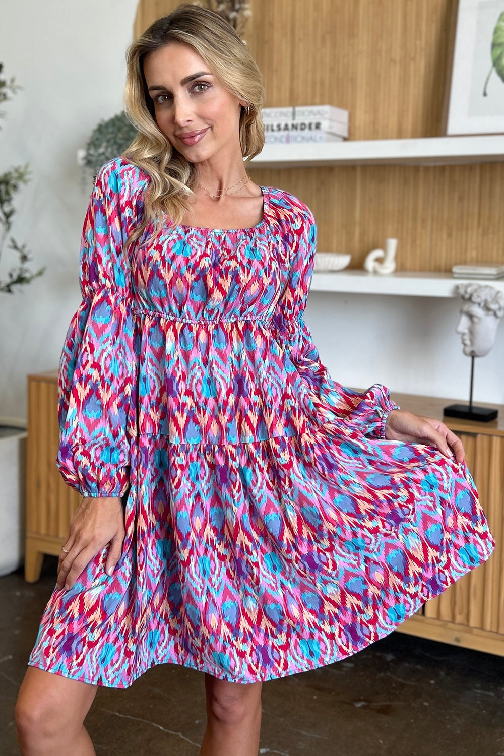 A model poses in a vibrant printed long sleeve dress with a square neckline, ruched waist, puff sleeves, tiered construction, and a flowing A-line silhouette.