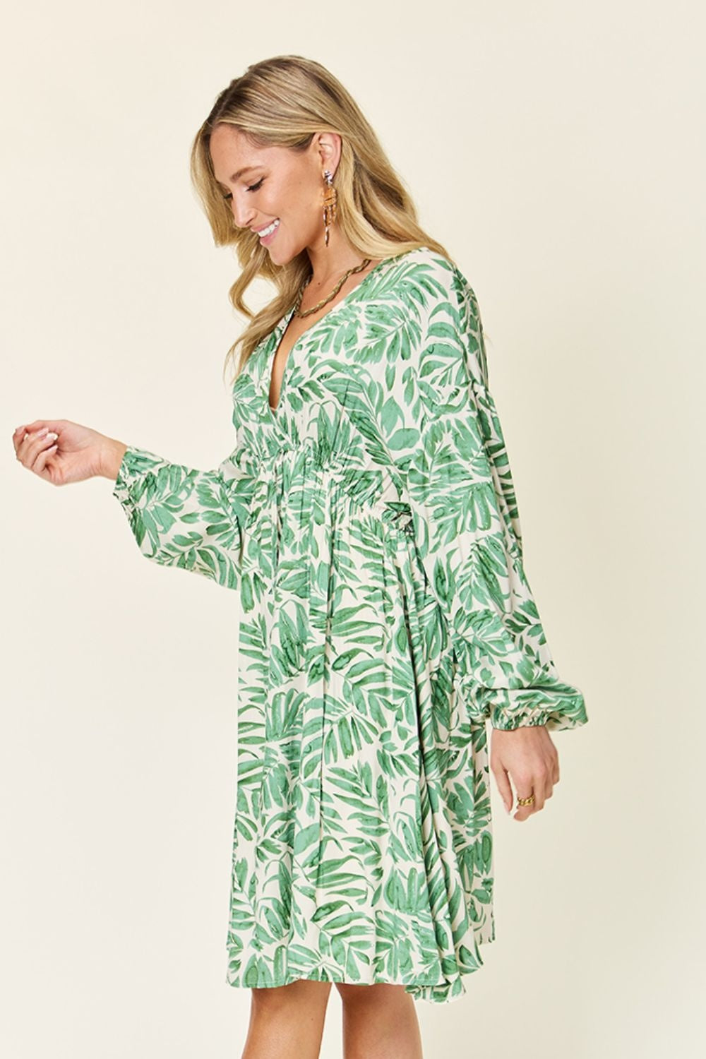 A model looks down while wearing a light green leafy-print dress with a V neckline, ruched waist, flowing silhouette, and voluminous balloon sleeves.