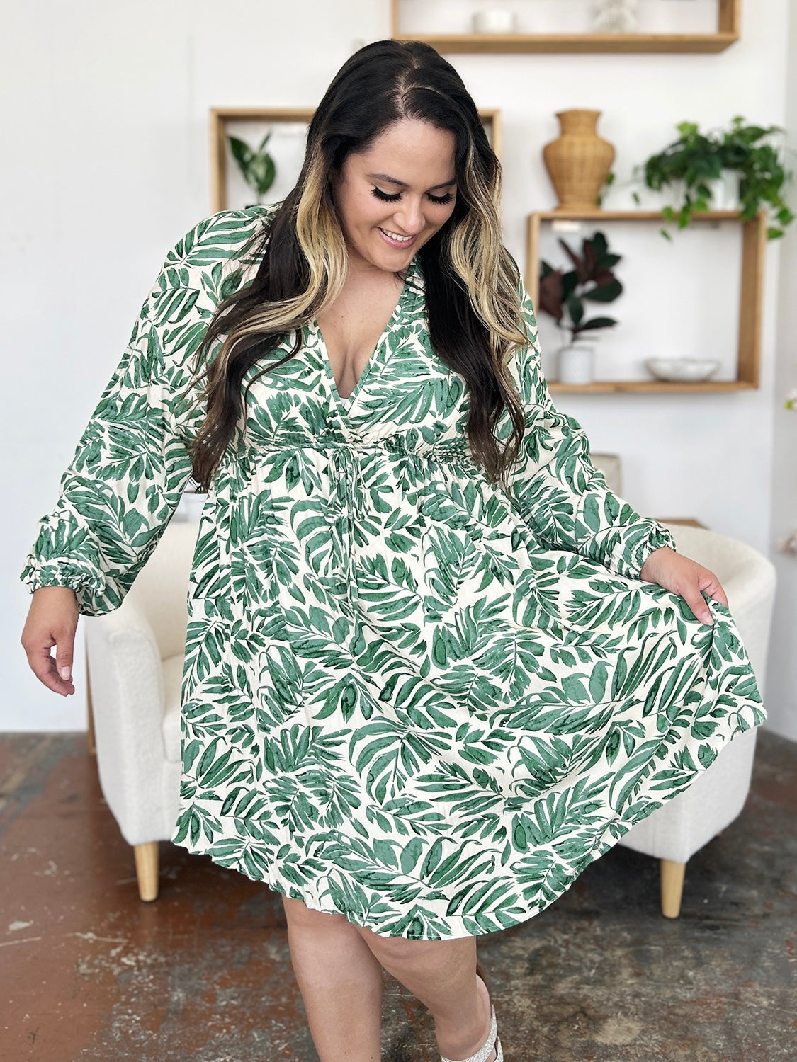 A model holds out the flowing skirt of a light green leafy printed dress with a V-neckline, ruched waist, and voluminous balloon sleeves.