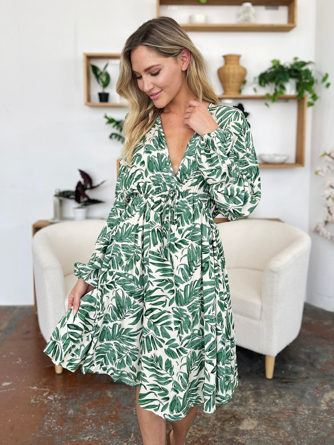 Model poses in a leafy green and cream printed dress with a V-neckline, ruched waist, voluminous balloon sleeves, and a flowing flared silhouette.