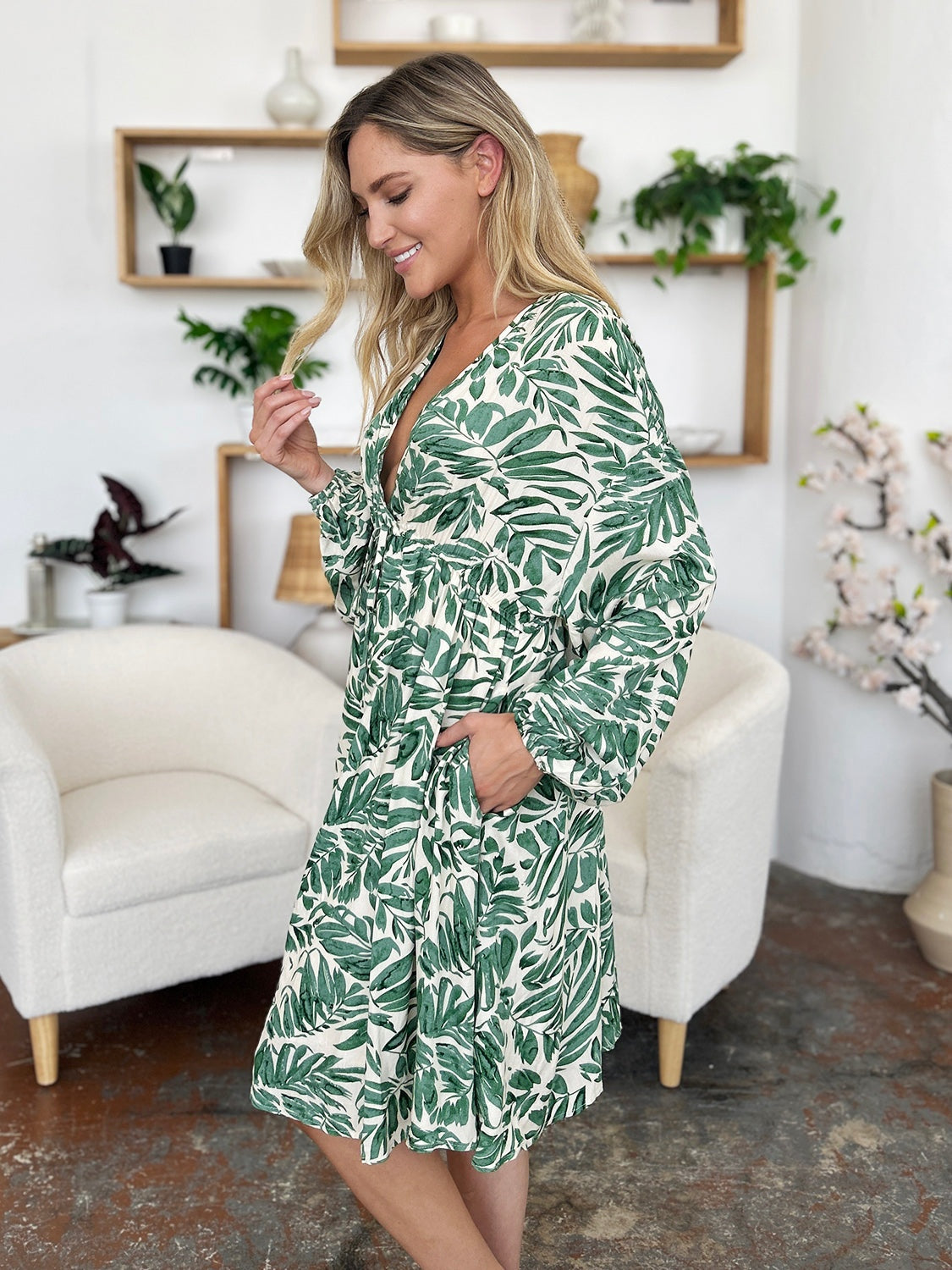 Model poses in a light green leafy-print dress with a V-neck, ruched waist, voluminous balloon sleeves, flowing skirt, and visible side pockets.