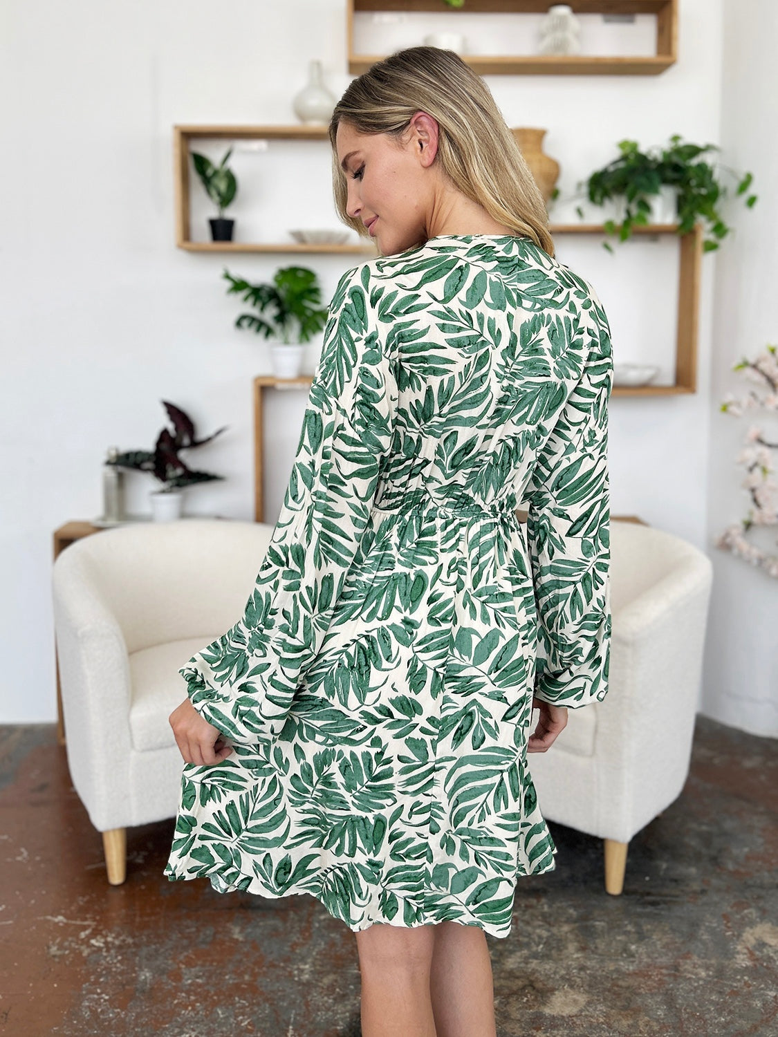 Model looks over her shoulder in a light green and white leafy-print dress with a ruched waist, voluminous balloon sleeves, and a softly flared skirt.
