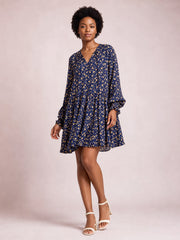 Model poses in a navy printed long-sleeve tiered dress with a flowy silhouette, gathered ruffle hem, and subtle tied neckline detail.