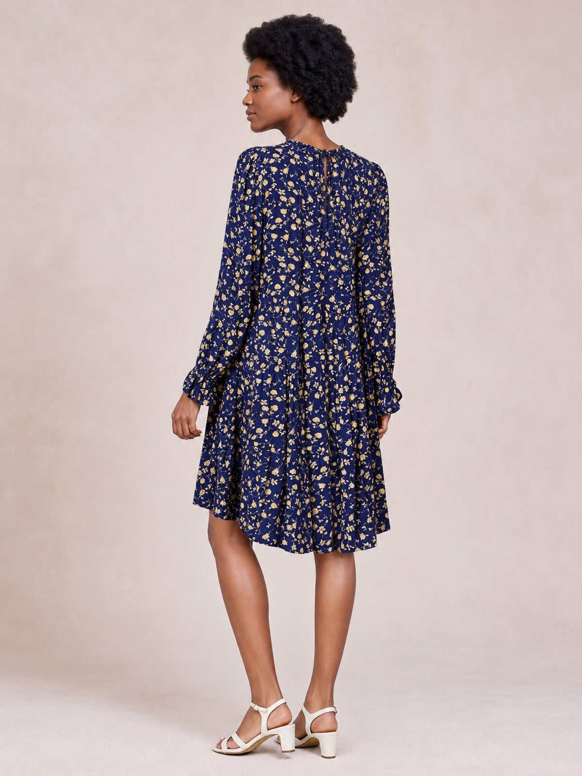 A model stands in a back-facing pose wearing a navy floral printed tiered dress with long sleeves, a tied neckline, gathered volume, and ruffled cuffs and hem.