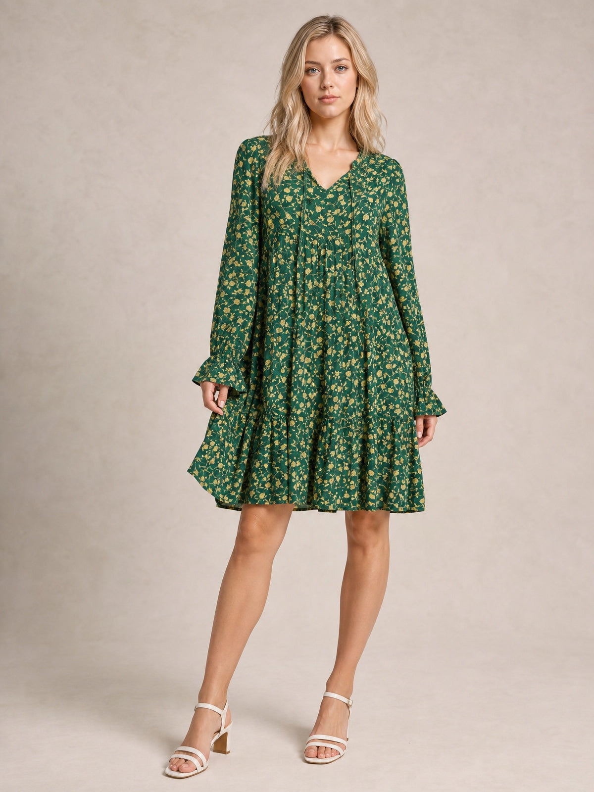A model poses in a green floral printed tiered dress with long sleeves, ruffled cuffs and hem, a gathered waist, and a flowy knee-length silhouette.