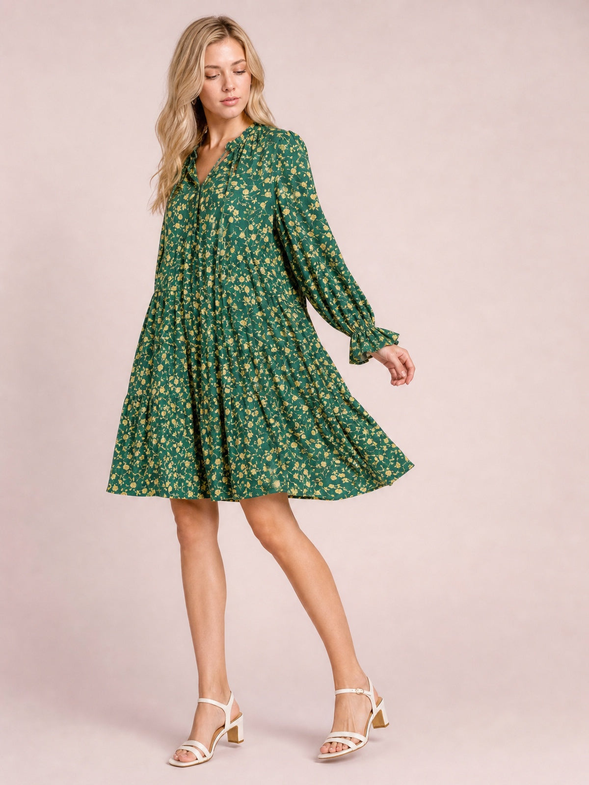 A model poses in a green floral printed tiered dress with long sleeves, a tied neckline, ruffled cuffs and hem, and a flowing A-line silhouette.