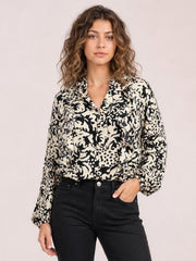 A model stands with one hand in her pocket wearing an opaque cream-and-black printed shirt with ruffle trim, button front, and voluminous balloon sleeves.
