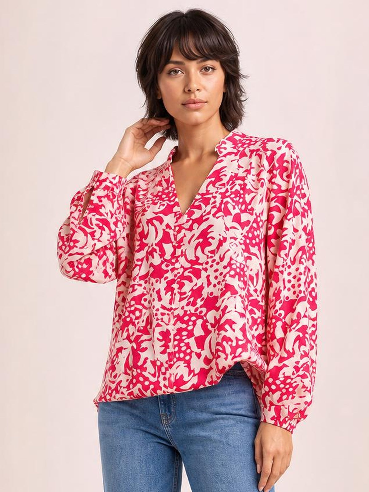 A model poses in a loose pink-and-white printed shirt with a ruffle-trimmed V-neck, voluminous balloon sleeves, and buttoned cuffs, lifting one hand to her neck.