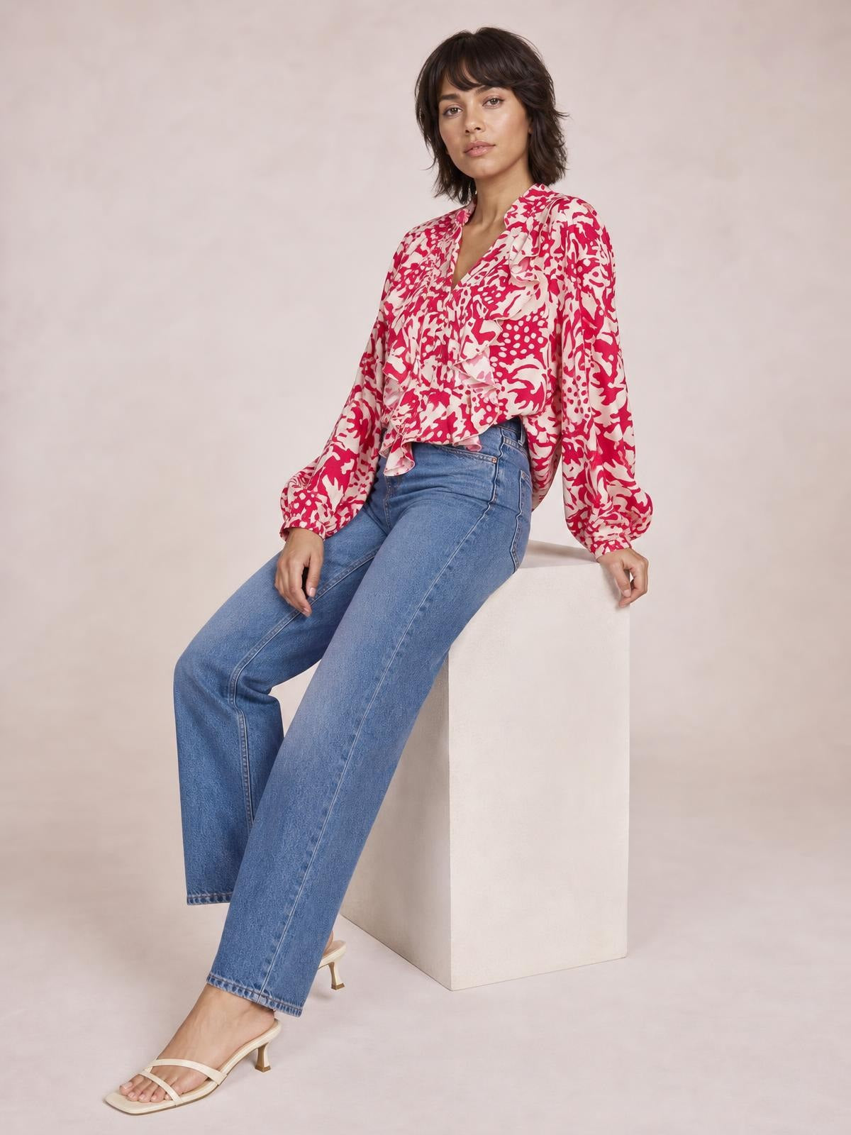 A seated model wears a red-and-white printed shirt with cascading ruffle trim, a buttoned front, and voluminous balloon sleeves.