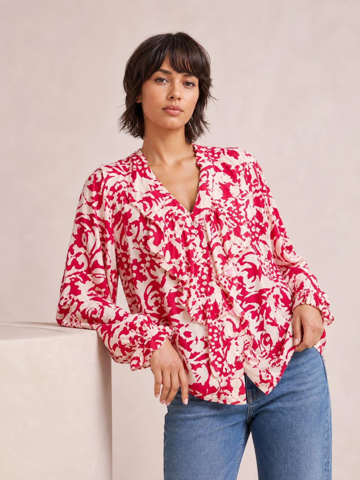 Model wears a pink and red printed shirt with ruffle trim, button front, V-neckline, and voluminous balloon sleeves with gathered cuffs.