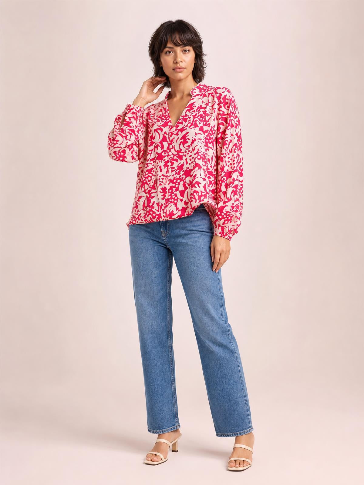 Model poses in a pink and white printed shirt with ruffle trim, a relaxed silhouette, V-neckline, and voluminous balloon sleeves with buttoned cuffs.