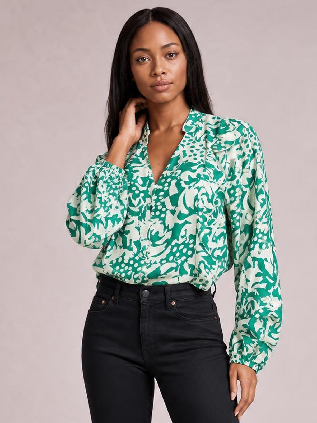 Model wears a green and white printed shirt with ruffle trim, button front, V neckline, and voluminous balloon sleeves while posing with one hand at her hair.