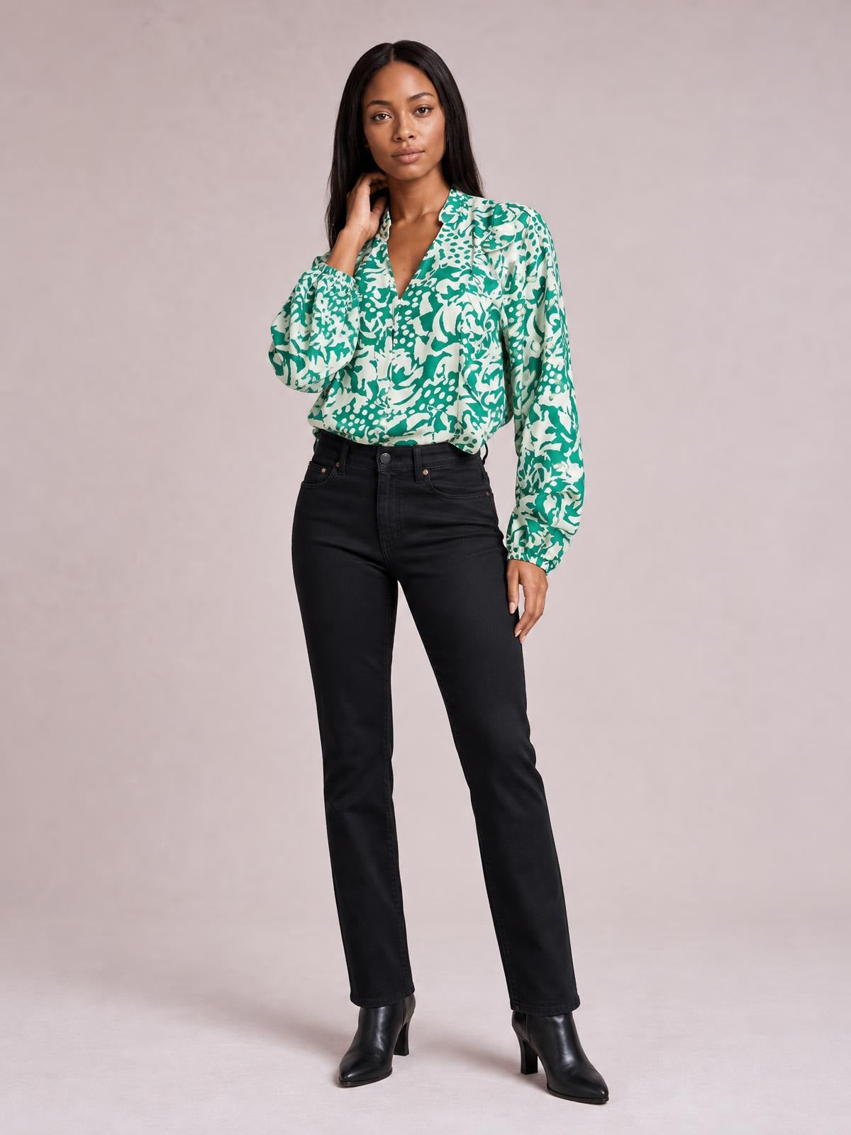 Model wearing a green and white printed shirt with ruffle trim, button front, and voluminous balloon sleeves, posing with one hand near her neck.
