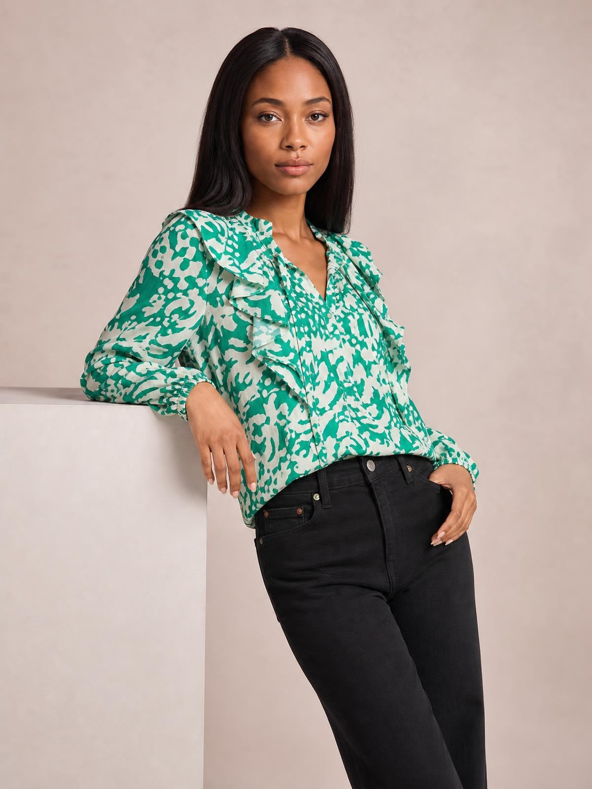 Model leans against a ledge wearing a green and white printed shirt with ruffle trim, buttoned front, and voluminous balloon sleeves.