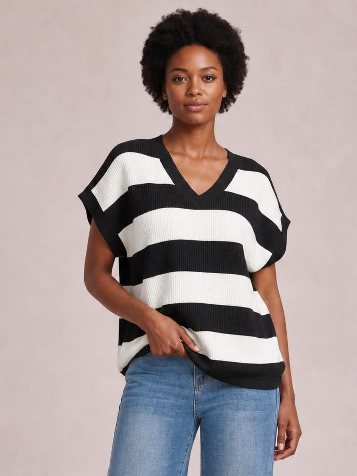 Model wearing a black-and-white striped V-neck short sleeve sweater with a relaxed silhouette, ribbed construction, and hands resting near the hem.
