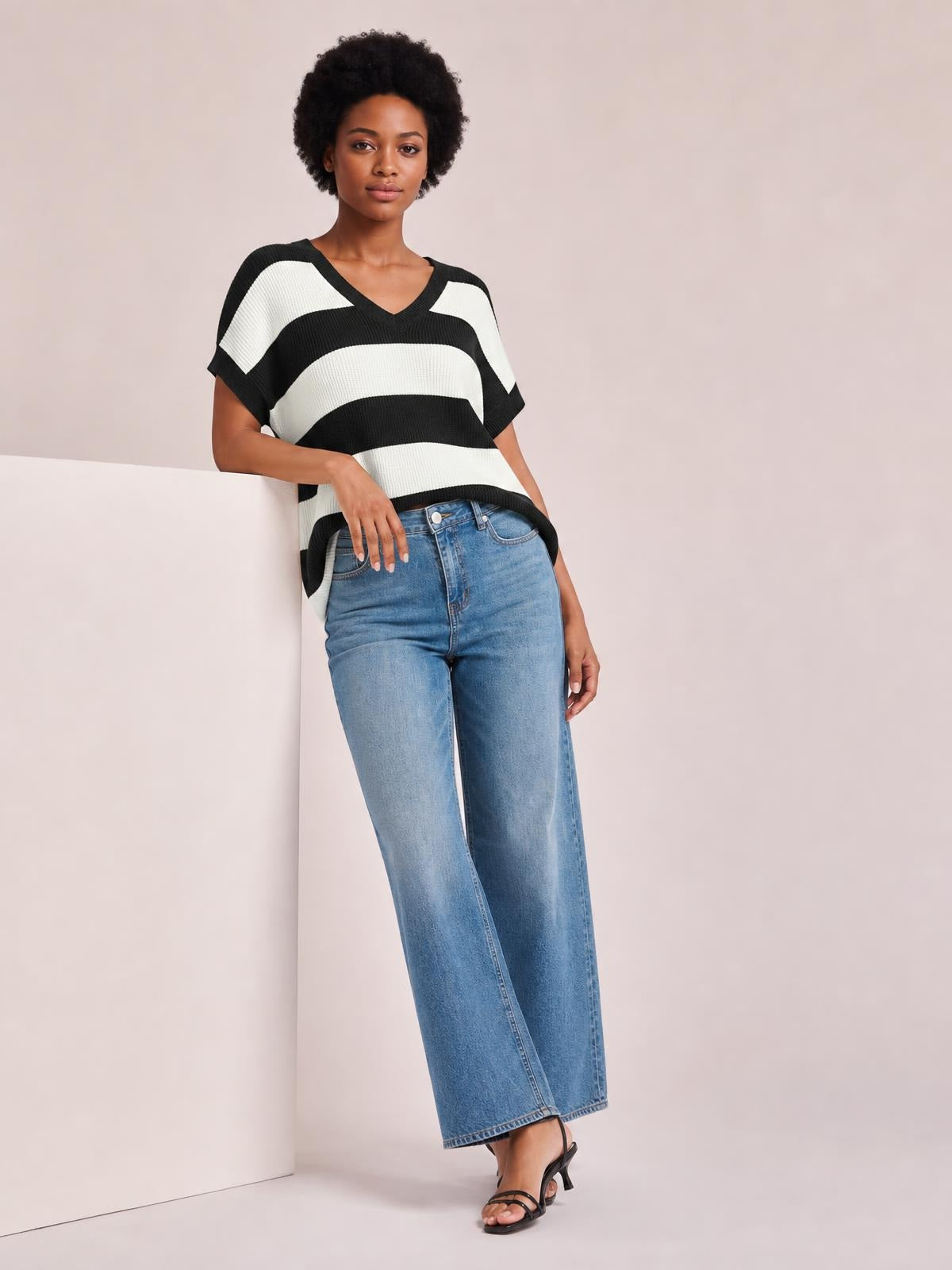 A model leans against a panel wearing a black-and-white striped V-neck short sleeve sweater with a relaxed, boxy silhouette and ribbed knit construction.