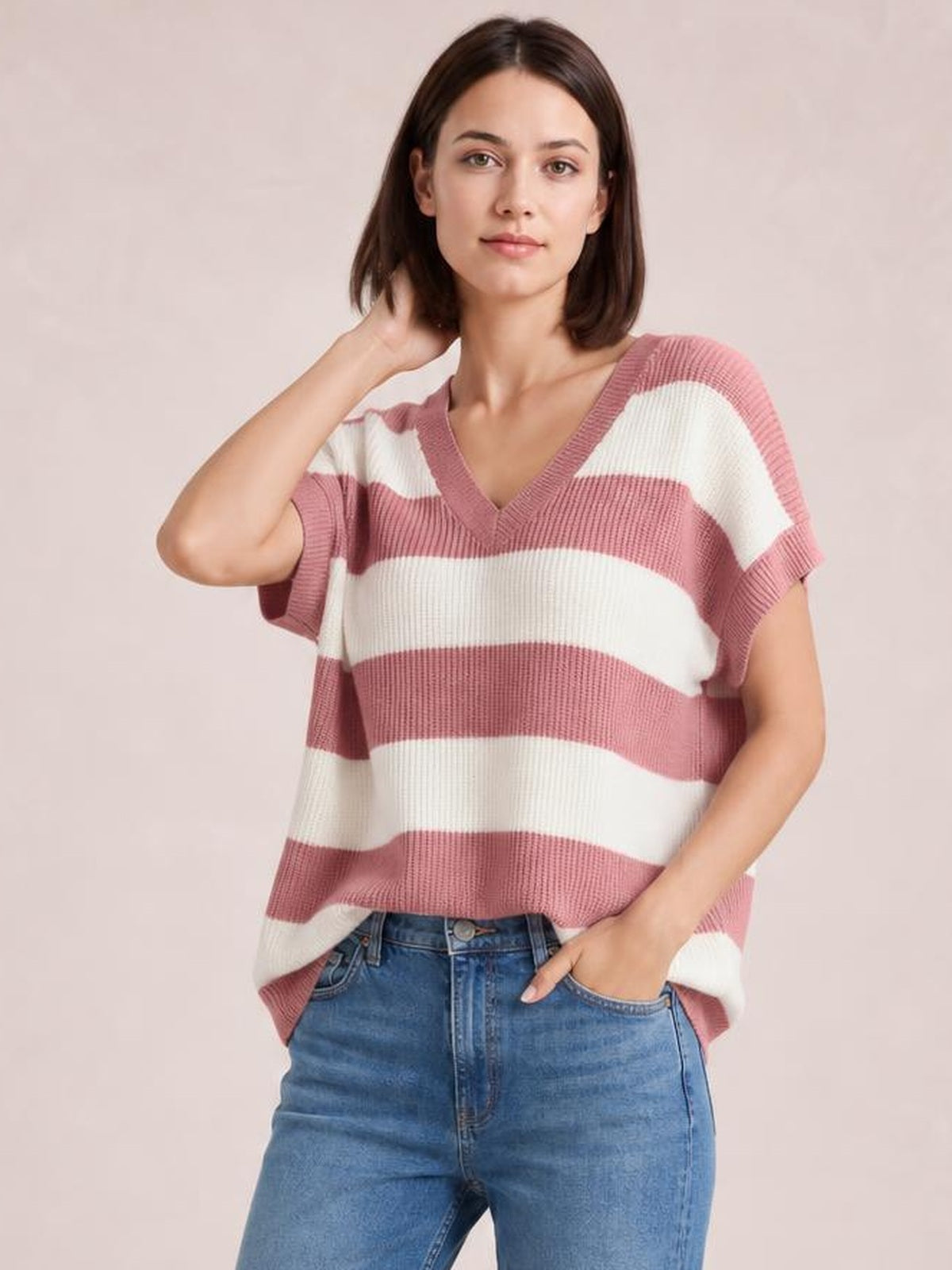 Model wearing a relaxed pink and white striped V-neck short sleeve sweater with ribbed trim, paired with jeans and one hand in a pocket.