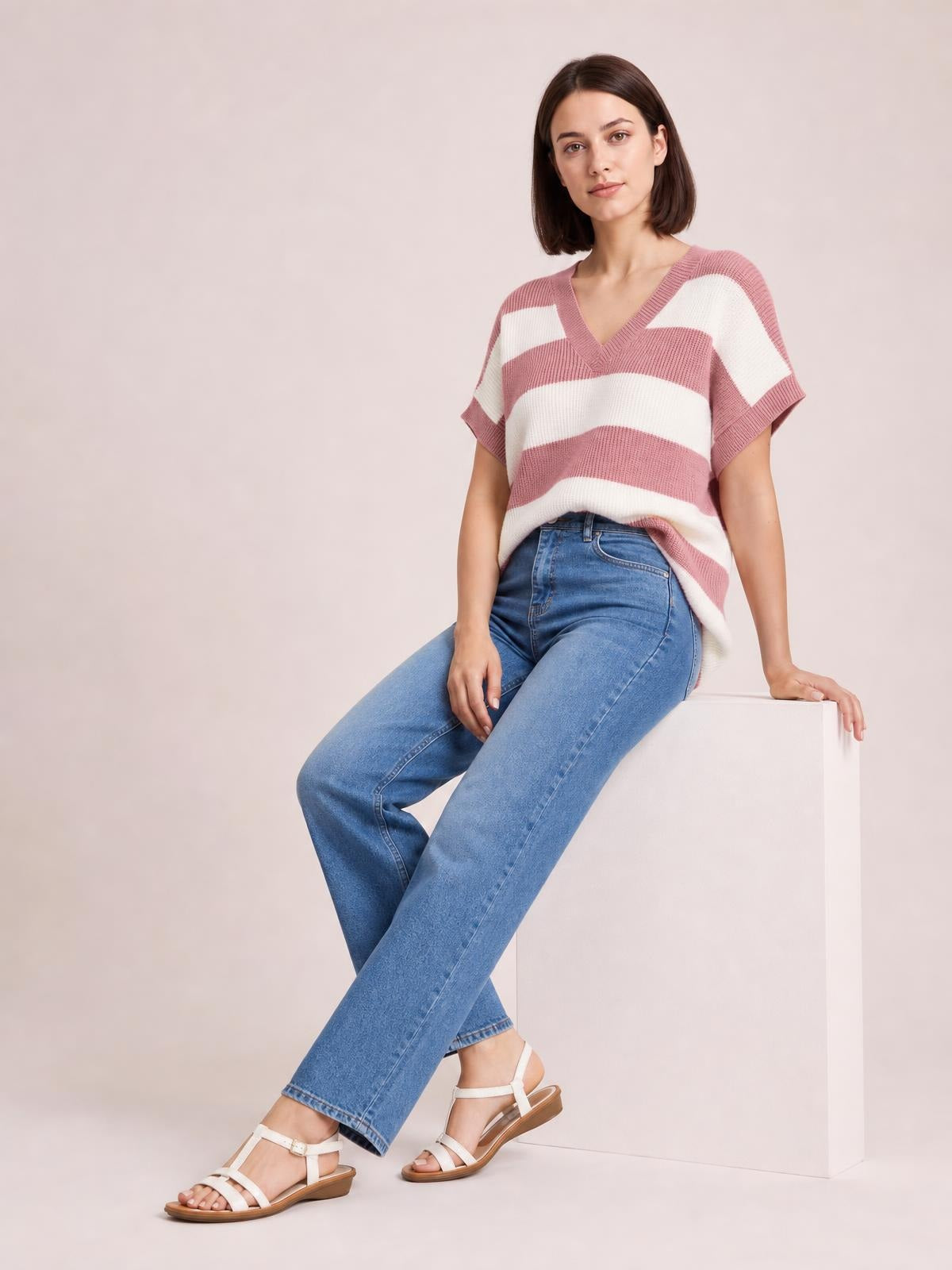 Model sits on a cube wearing a pink and white striped V-neck short sleeve sweater with ribbed trim and a relaxed silhouette.