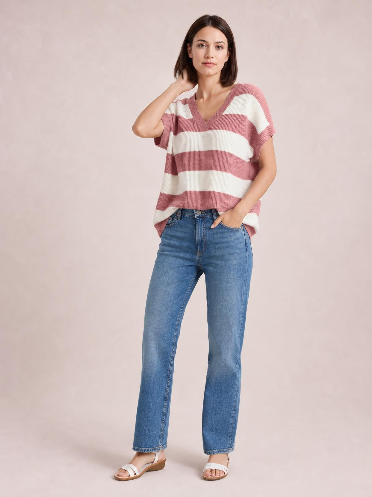 Model wearing a pink and cream striped V-neck short-sleeve sweater with ribbed trim and a relaxed silhouette, posing with one hand in her jeans pocket.