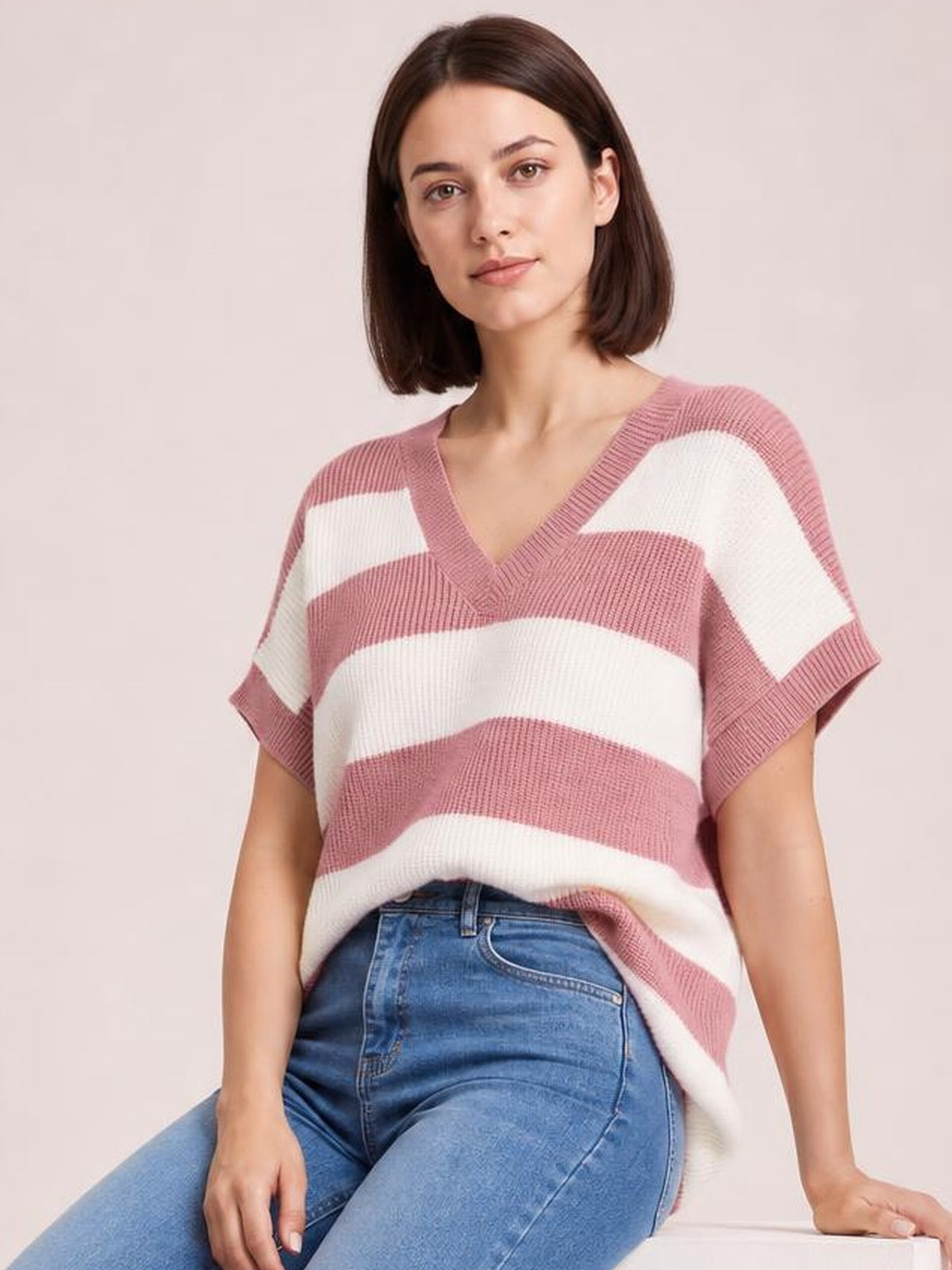Model seated in a striped pink and white V-neck short sleeve sweater with ribbed trim, relaxed silhouette, and wide horizontal bands.