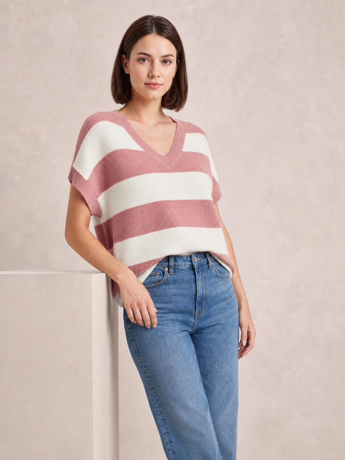 A model leans casually in a relaxed pink-and-cream striped V-neck short sleeve sweater with ribbed trim and a loose silhouette.