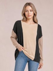 Model wearing a relaxed round-neck long-sleeve T-shirt with beige ribbed texture, black contrast panels, and a curved hem while posing with one hand at the hem.