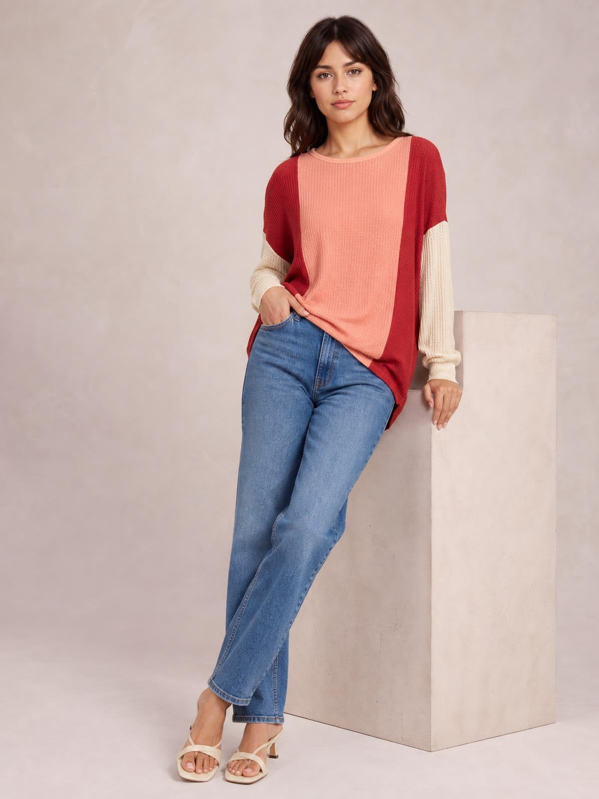 Model leans against a block wearing a relaxed texture contrast round neck long sleeve T-shirt in coral, red, and cream with a color-blocked silhouette.