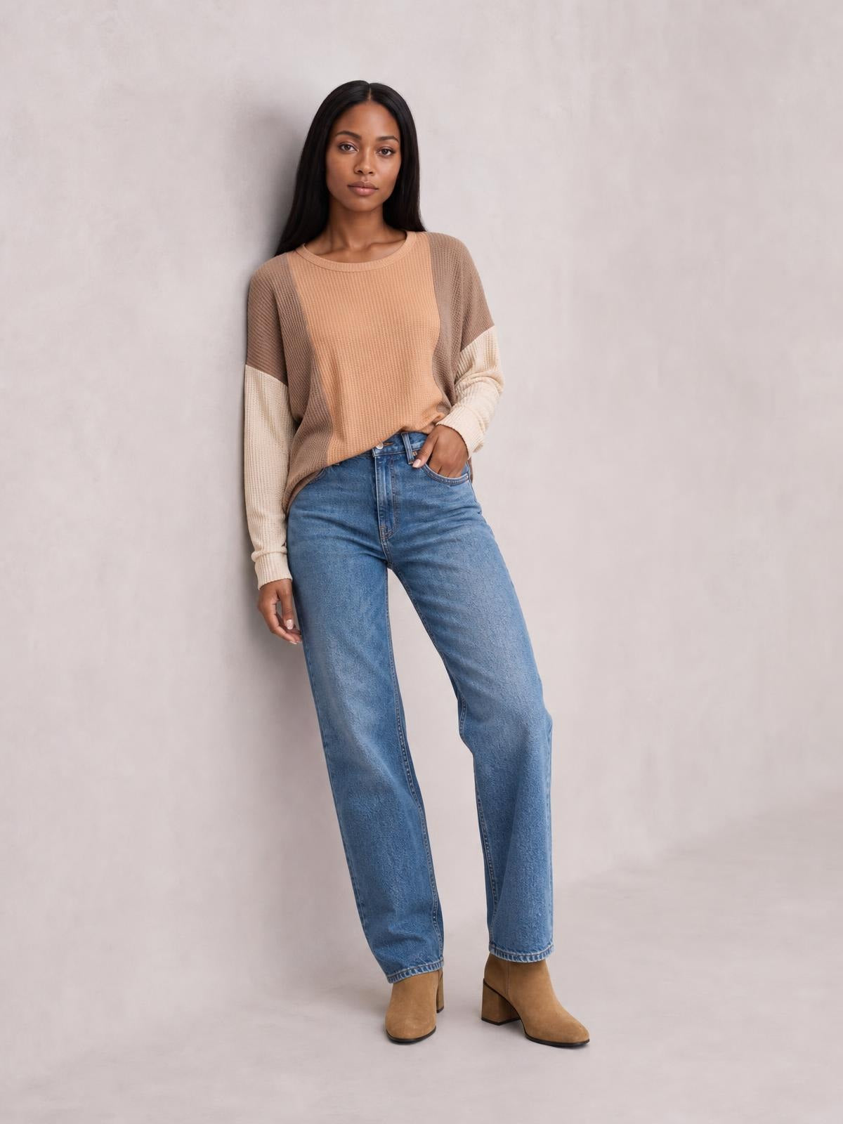 A model leans casually in a tan textured long-sleeve T-shirt with a round neck, contrasting brown shoulders and cream sleeve panels, in a relaxed silhouette.