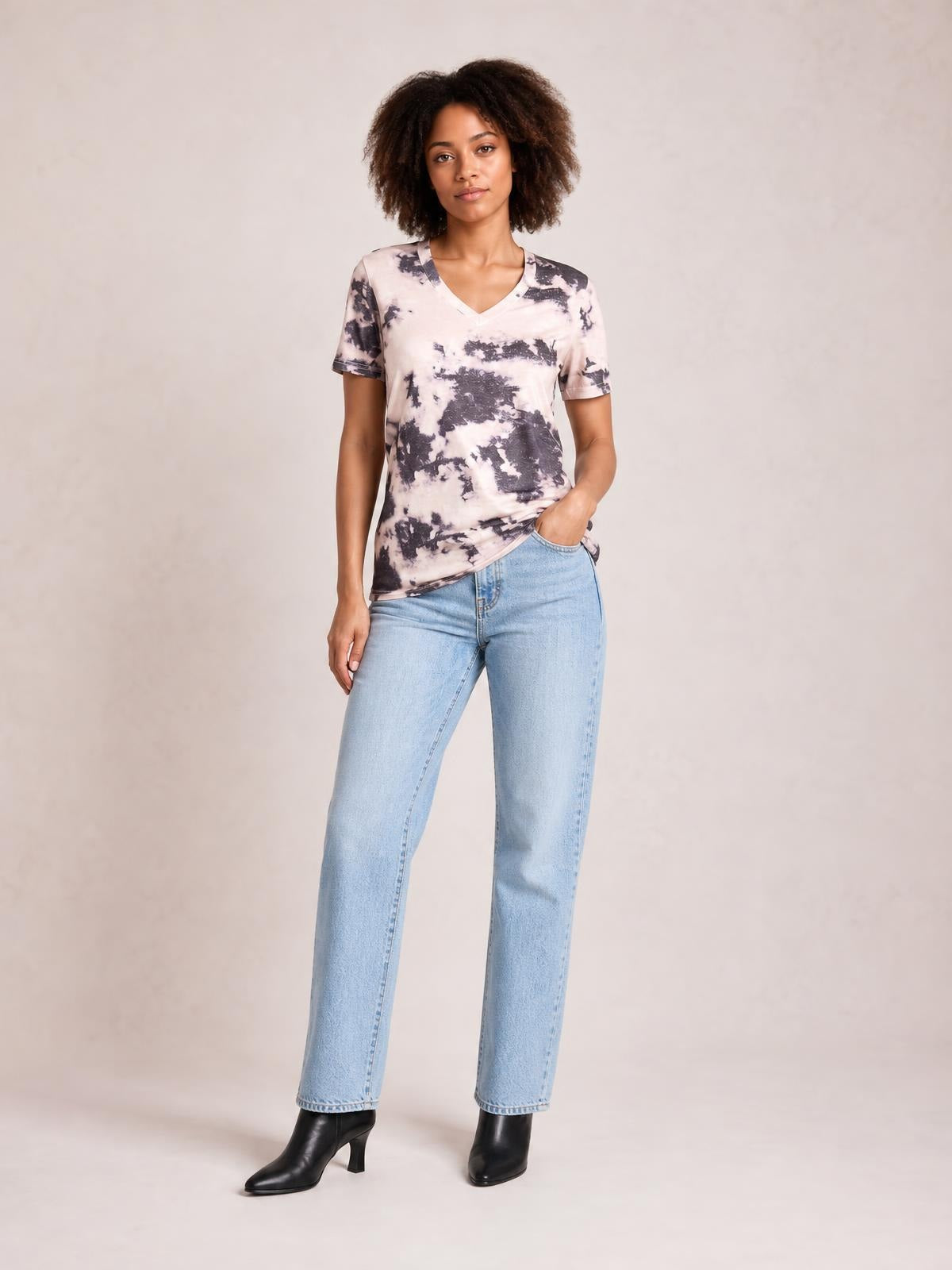 Model wearing an opaque pink and charcoal tie-dye V-neck T-shirt with short sleeves and a relaxed straight silhouette, standing with one hand in her jeans pocket.