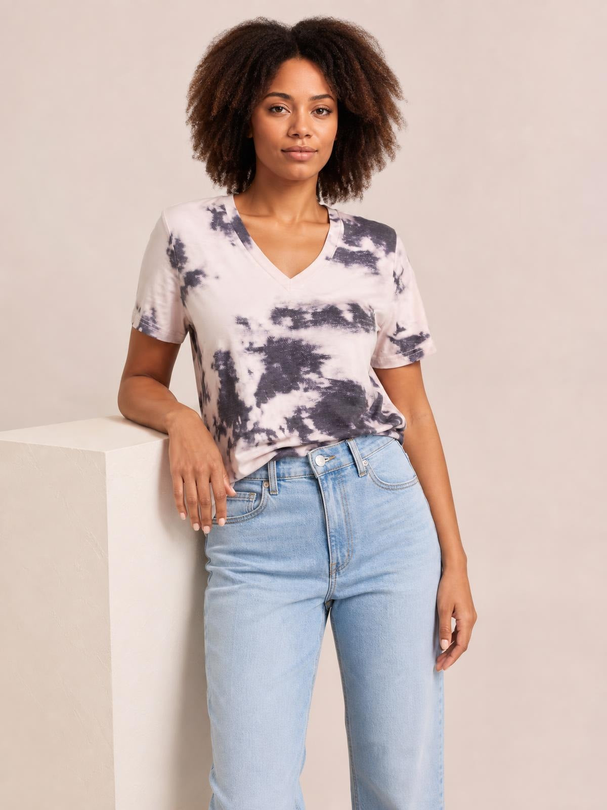 Model poses in a black-and-white tie-dye V-neck T-shirt with short sleeves and a relaxed silhouette, leaning one arm on a pedestal.