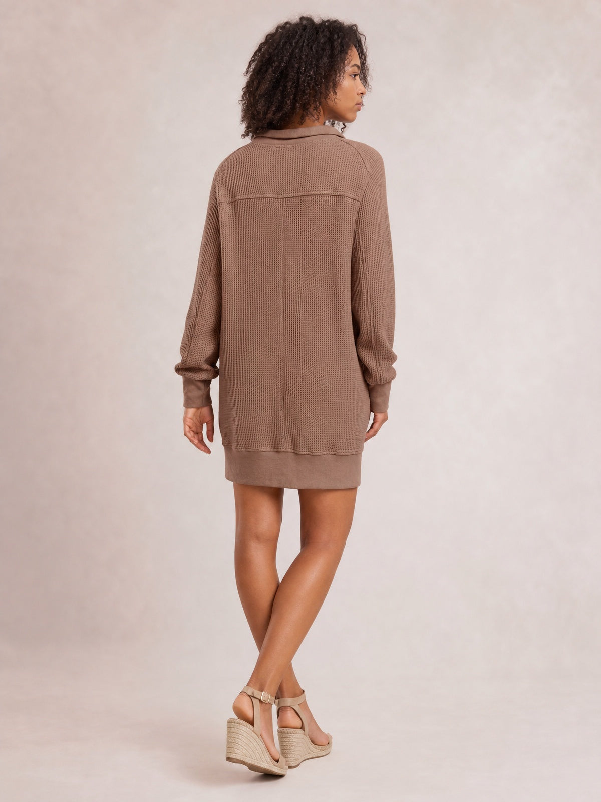 Model looks over her shoulder in a taupe waffle-knit long-sleeve mini dress with a relaxed straight silhouette, textured panels, and ribbed cuffs and hem.