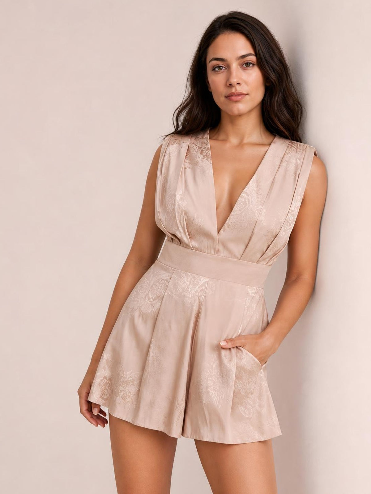A model leans in a blush pink draped romper with a V-neck, sleeveless pleated shoulder pad detail, wide waistband, and flowing floral-jacquard shorts.
