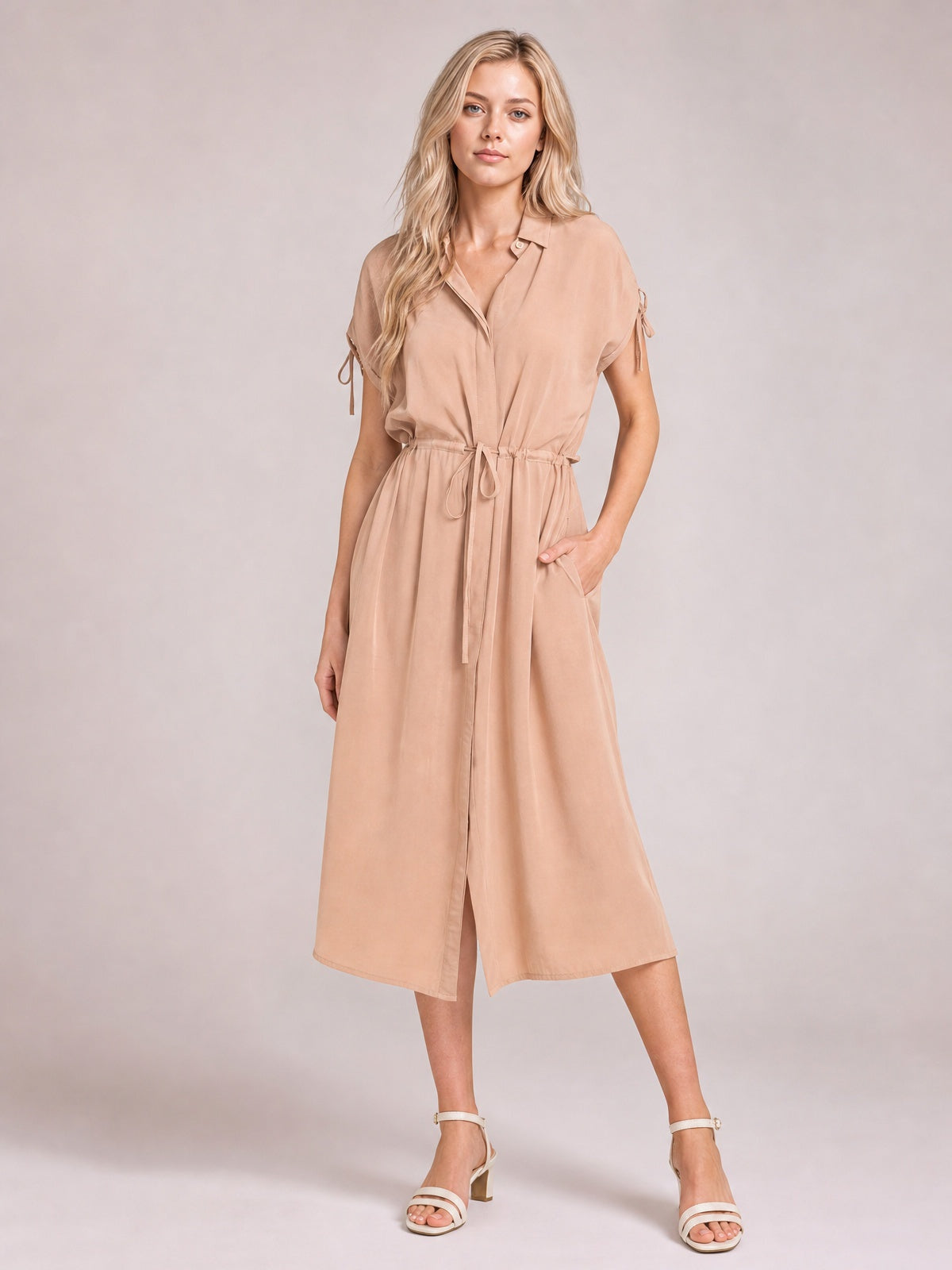 A model poses in a tan collared neck short-sleeve midi dress with a buttoned front, drawstring waist, side pockets, and softly flared silhouette.