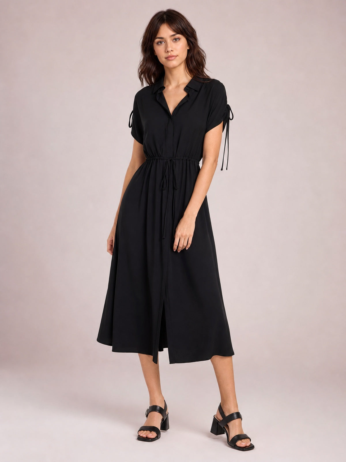 Model poses in a black collared neck midi dress with short sleeves, buttoned front, drawstring waist, and a fluid, softly flared skirt.