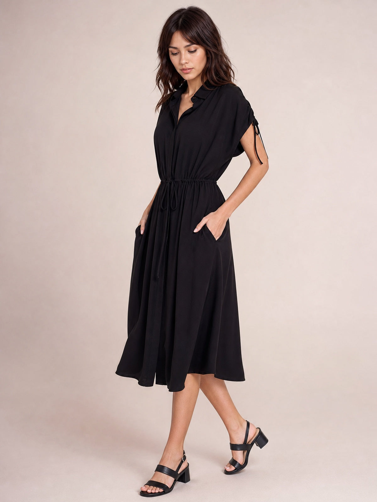 Model poses in a black collared neck midi dress with short sleeves, buttoned front, drawstring waist, pockets, and a softly flared silhouette.