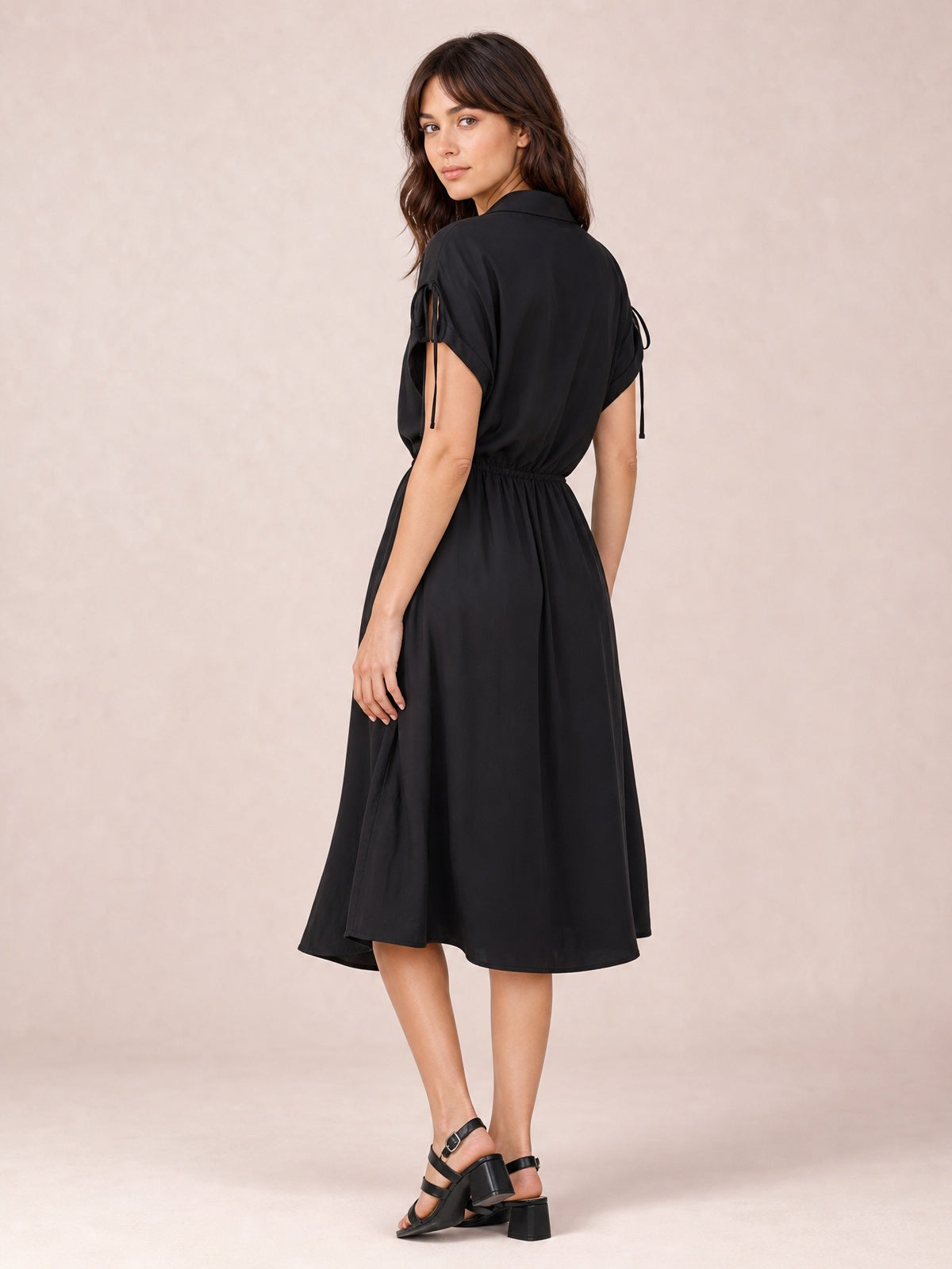 A model looks over her shoulder in a black collared neck short sleeve midi dress with a gathered drawstring waist and softly flared skirt.
