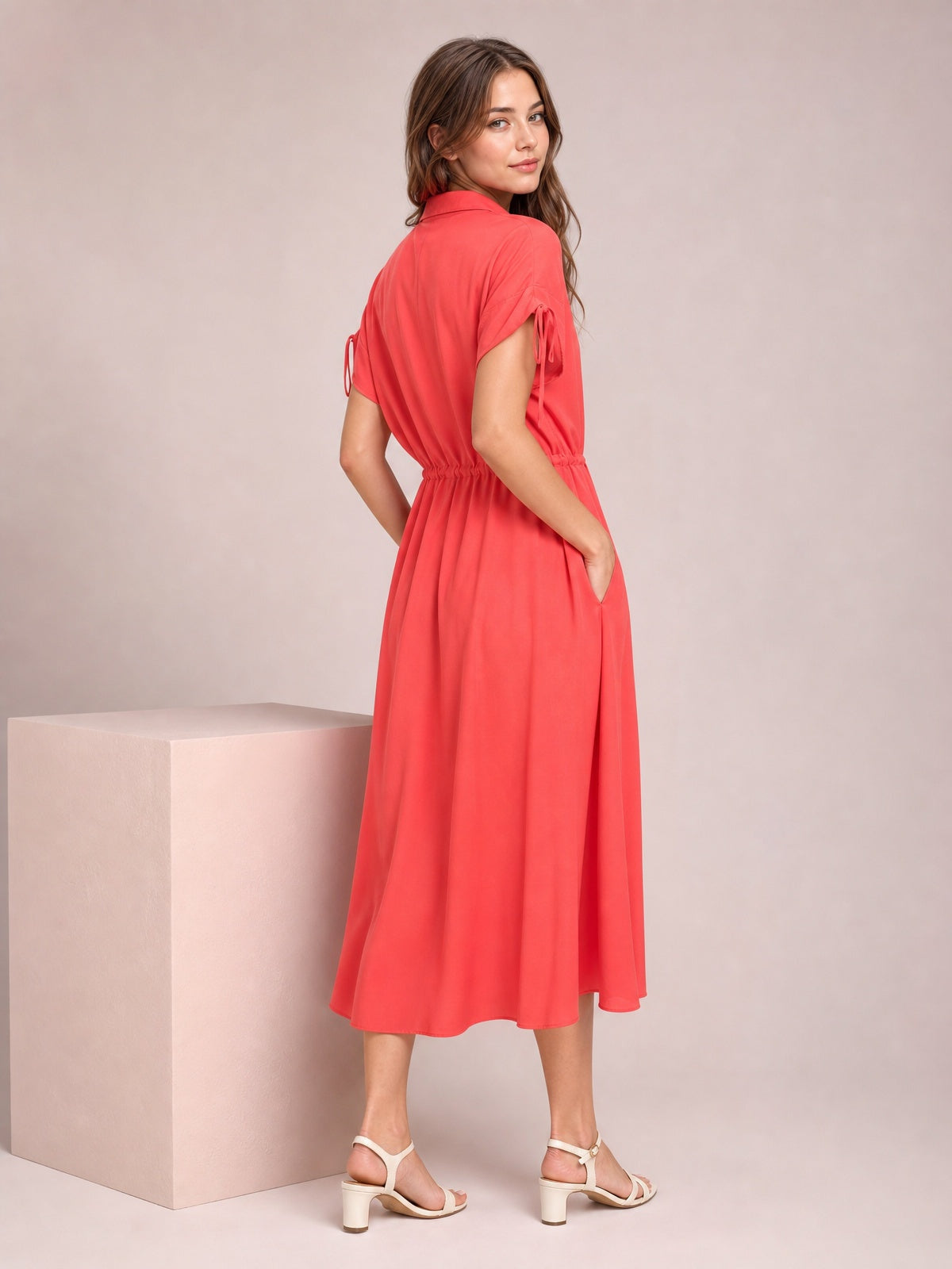 A model poses in a coral drawstring collared neck midi dress with short sleeves, a gathered waist, and a fluid, softly flared skirt.