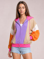 Model stands with one hand on her hip wearing a color-block zip-up hoodie with a purple front, tan panels, orange sleeves, pink drawstrings, and a ribbed hem.