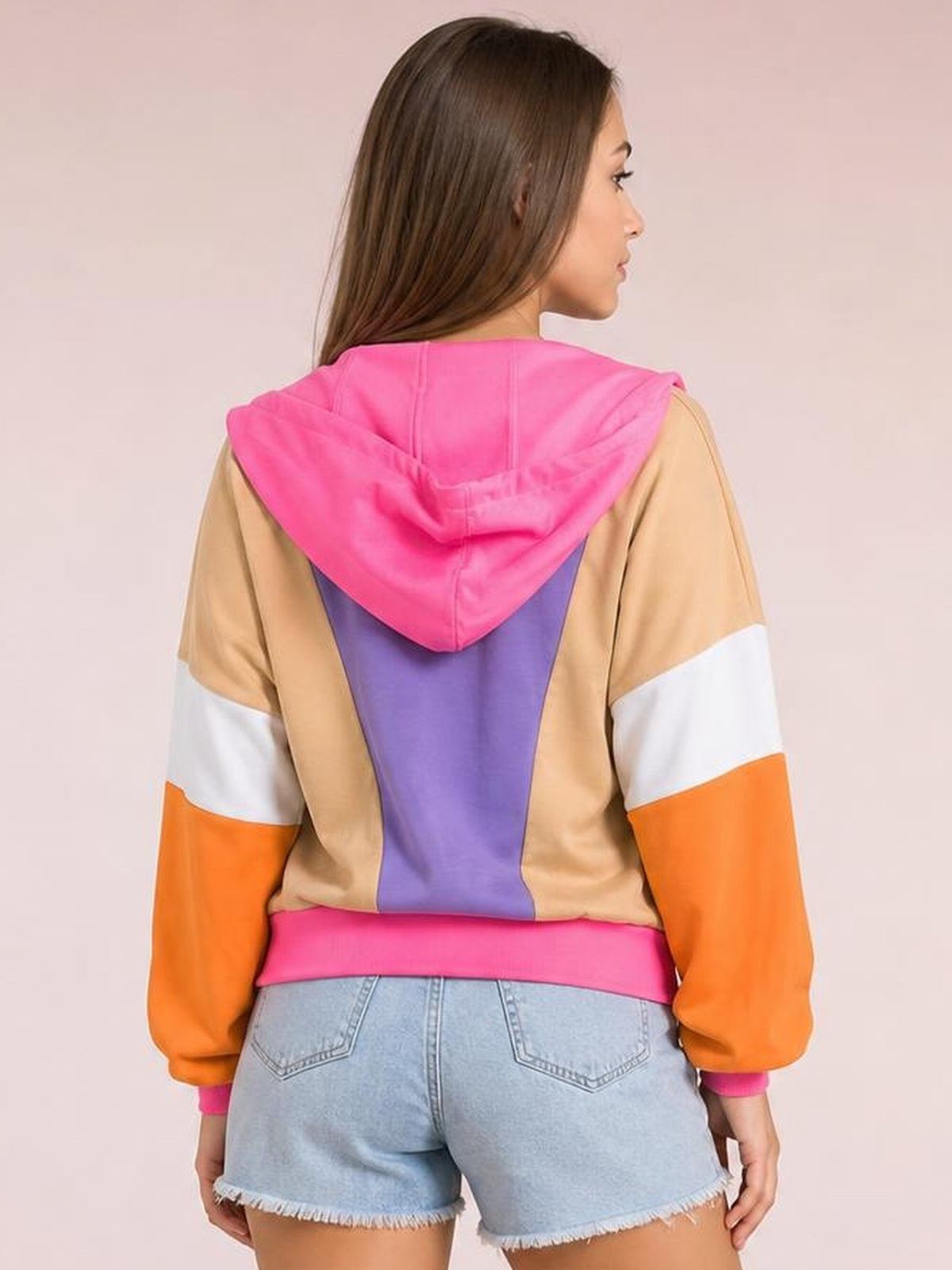 A model faces away wearing a color block long-sleeve hoodie with a bright pink hood, tan body, purple back panel, and orange-and-white sleeves.