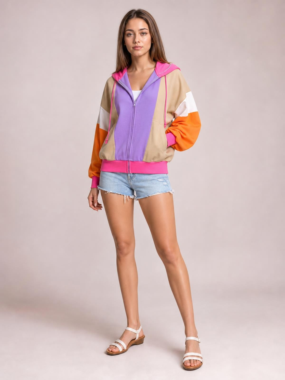 Model poses in a color block zip-up long-sleeve hoodie with a purple front, beige panels, orange sleeves, pink trim, drawstring hood, and ribbed hem.