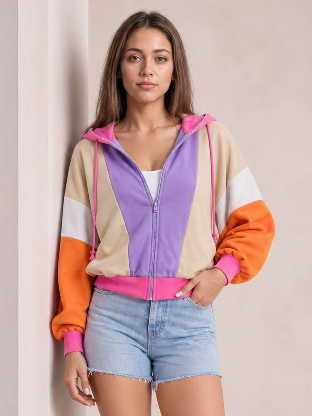 Model poses with one hand in her pocket wearing a color-block zip-up hoodie with a purple front, tan body, orange sleeves, pink drawstrings, and long sleeves.