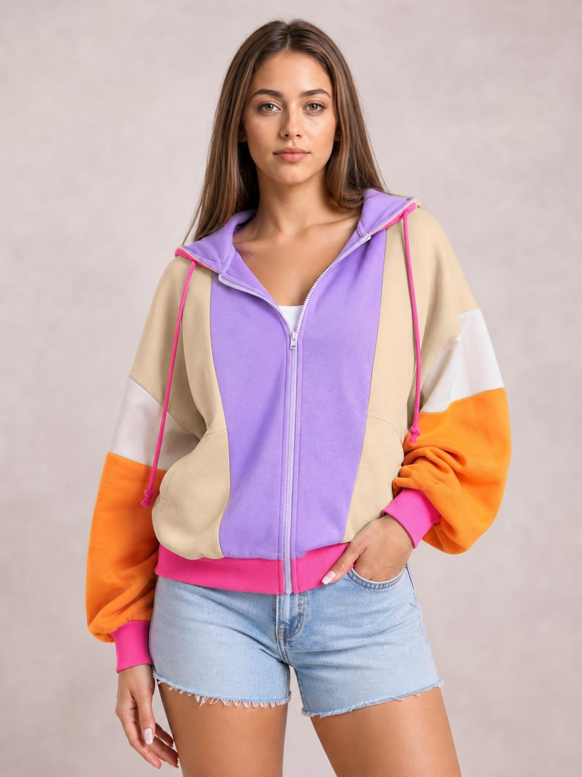 Model wearing a color block zip-up long-sleeve hoodie with a purple front, beige panels, orange sleeves, pink drawstrings, and a fitted silhouette.